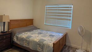 Quarto com cama, mesa de cabeceira, abajur, ventoinha no teto, janela com persianas e ventilador.