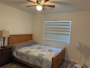 Un dormitorio con cama, mesita de noche, lámpara, ventilador de techo, ventana con persianas y ventilador de pie.