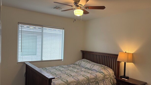 Quarto com cama, mesa de cabeceira, luminária, ventilador de teto e janela com persianas.