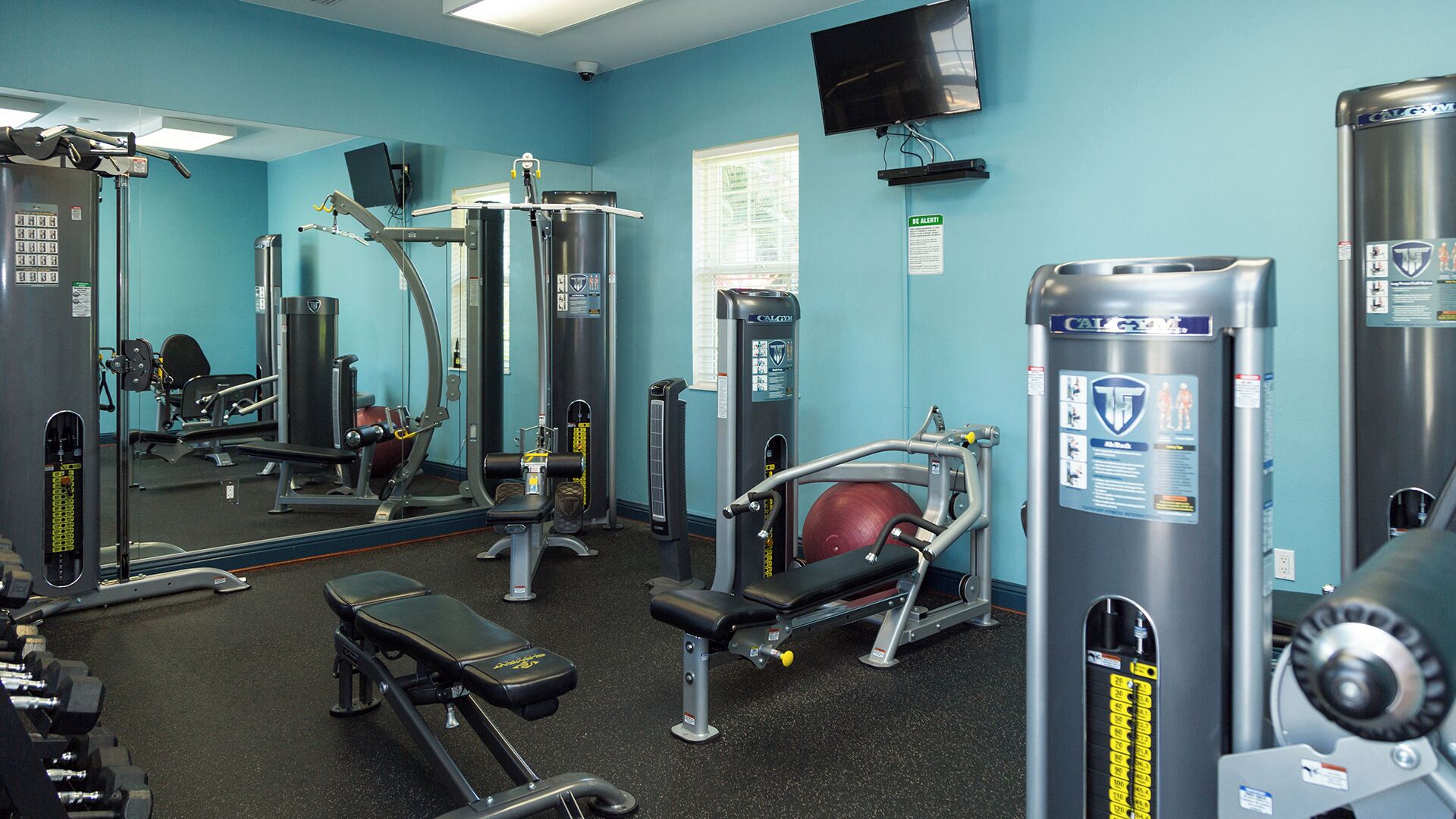A gym with various exercise equipment, mirrors, and a television on the wall.