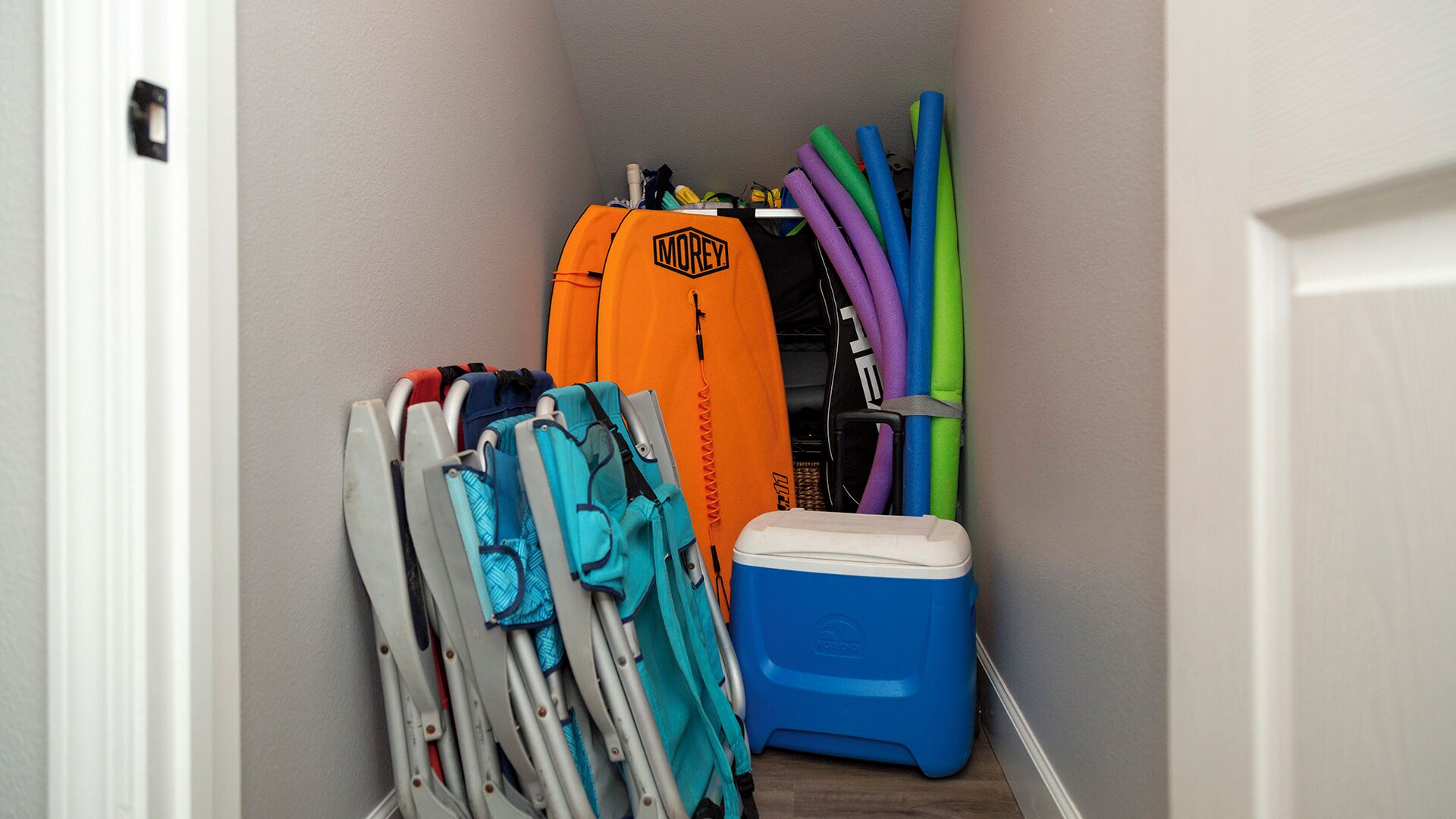 A narrow hallway with a white door on the left, a blue cooler, and various items including a blue and orange bag, a white and blue chair, and colorful foam rollers.
