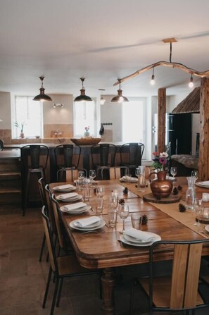 Uma sala de jantar com uma mesa de madeira comprida posta para refeição, rodeada por cadeiras, e iluminação pendente acima.