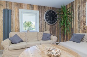 Quarto com parede de madeira, um relógio redondo grande, um sofá bege, mesa de centro de madeira e um vaso com planta.