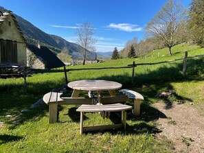 Ein Holztisch mit Bänken in einer grünen Wiese, umgeben von einem Holzzaun, mit Bergen im Hintergrund.