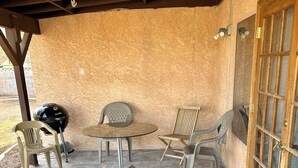 Uma varanda coberta com piso de pedra, uma mesa redonda, duas cadeiras de plástico, uma churrasqueira e uma porta de madeira com painéis de vidro.