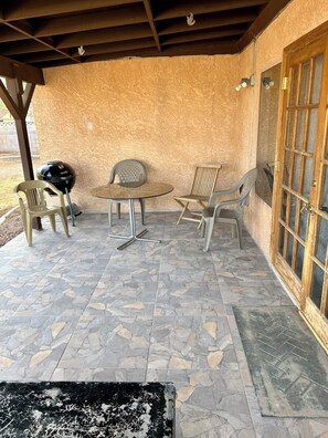 Un patio techado con piso de piedra, una mesa redonda, dos sillas de plástico, una parrilla y una puerta de madera con paneles de vidrio.