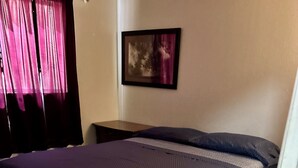 Uma cama de solteiro com colcha azul escura e estrutura de madeira, uma mesa de cabeceira, um quadro na parede e uma porta à direita.
