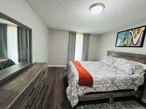 Un dormitorio con una cama, un espejo grande, un cuadro y una ventana con cortinas.