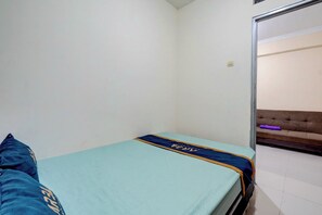 Quarto pequeno com uma cama de solteiro, parede branca simples, porta visível e um sofá marrom ao fundo.