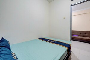 Una habitación pequeña con cama individual, pared blanca lisa, una puerta visible y un sofá marrón al fondo.