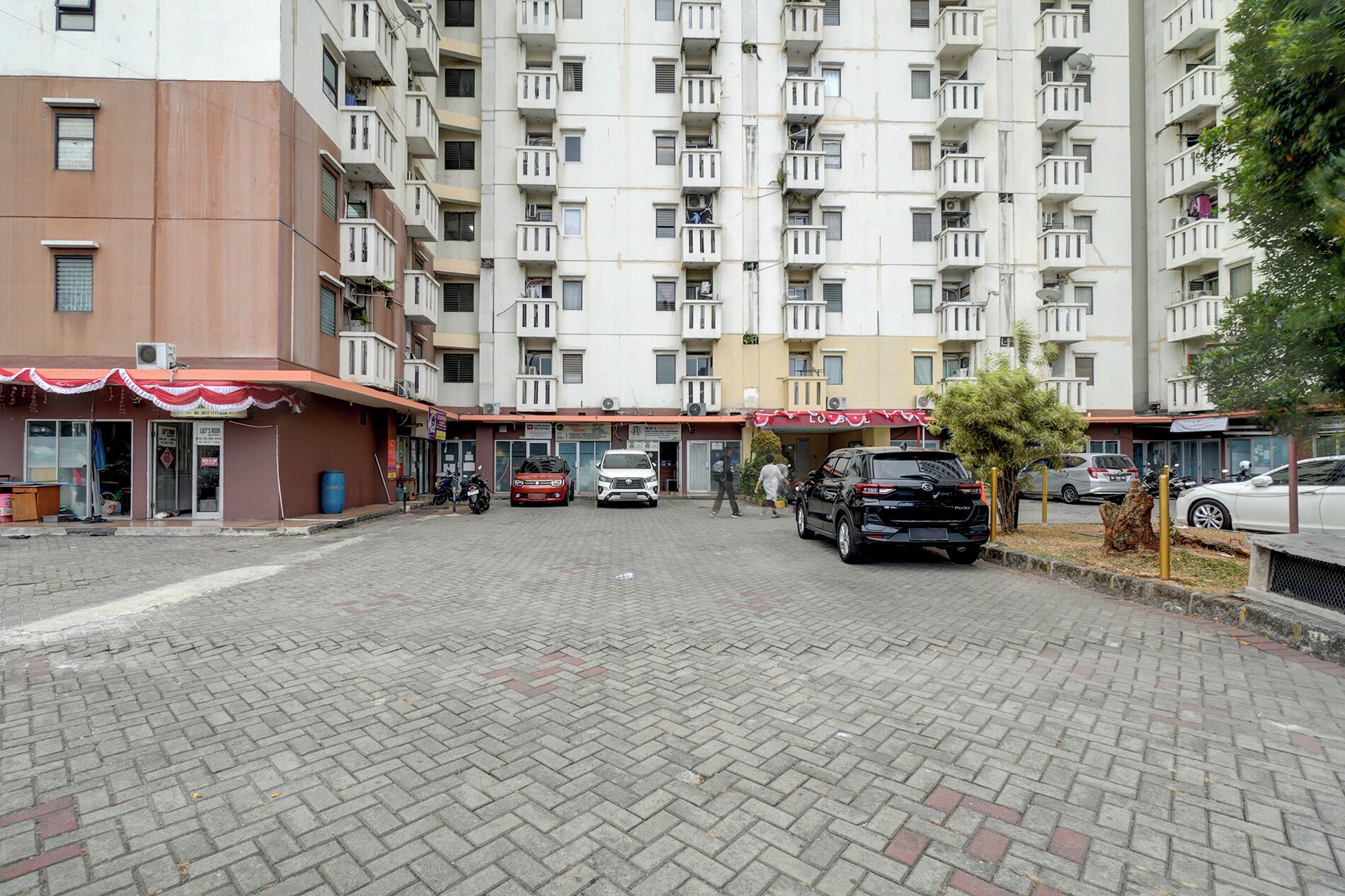 Un edificio residencial de varios pisos con un acceso pavimentado, varios coches aparcados y algunas personas caminando.