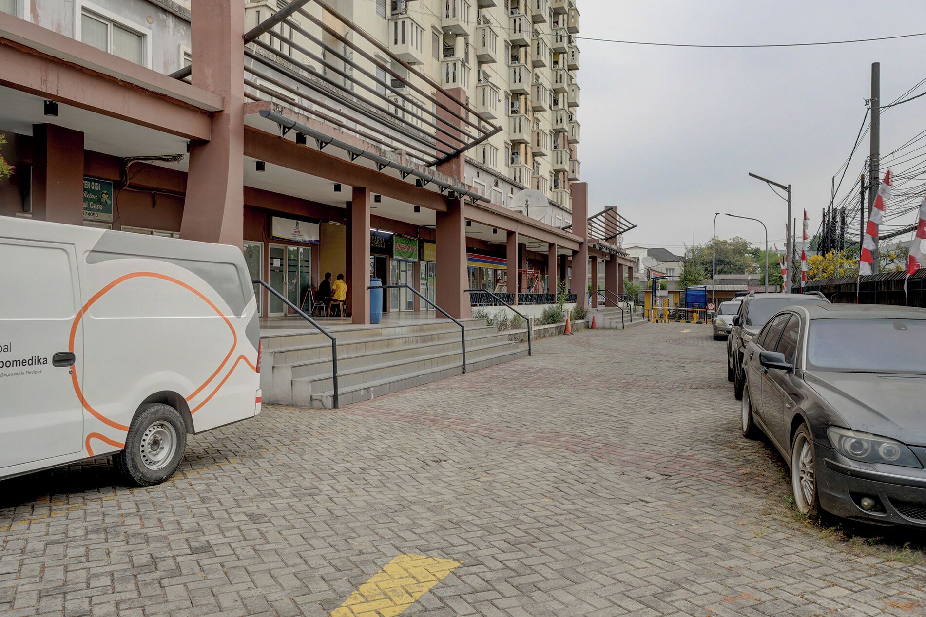 Un área pavimentada con una furgoneta estacionada a la izquierda, un edificio de varios pisos al fondo y un automóvil a la derecha.