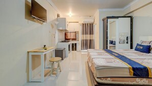 A hotel room with a bed, a small table, a chair, a TV mounted on the wall, and a bathroom visible through an open door. - Apartemen Cibubur Village KaisarProperty (Kota Depok)
