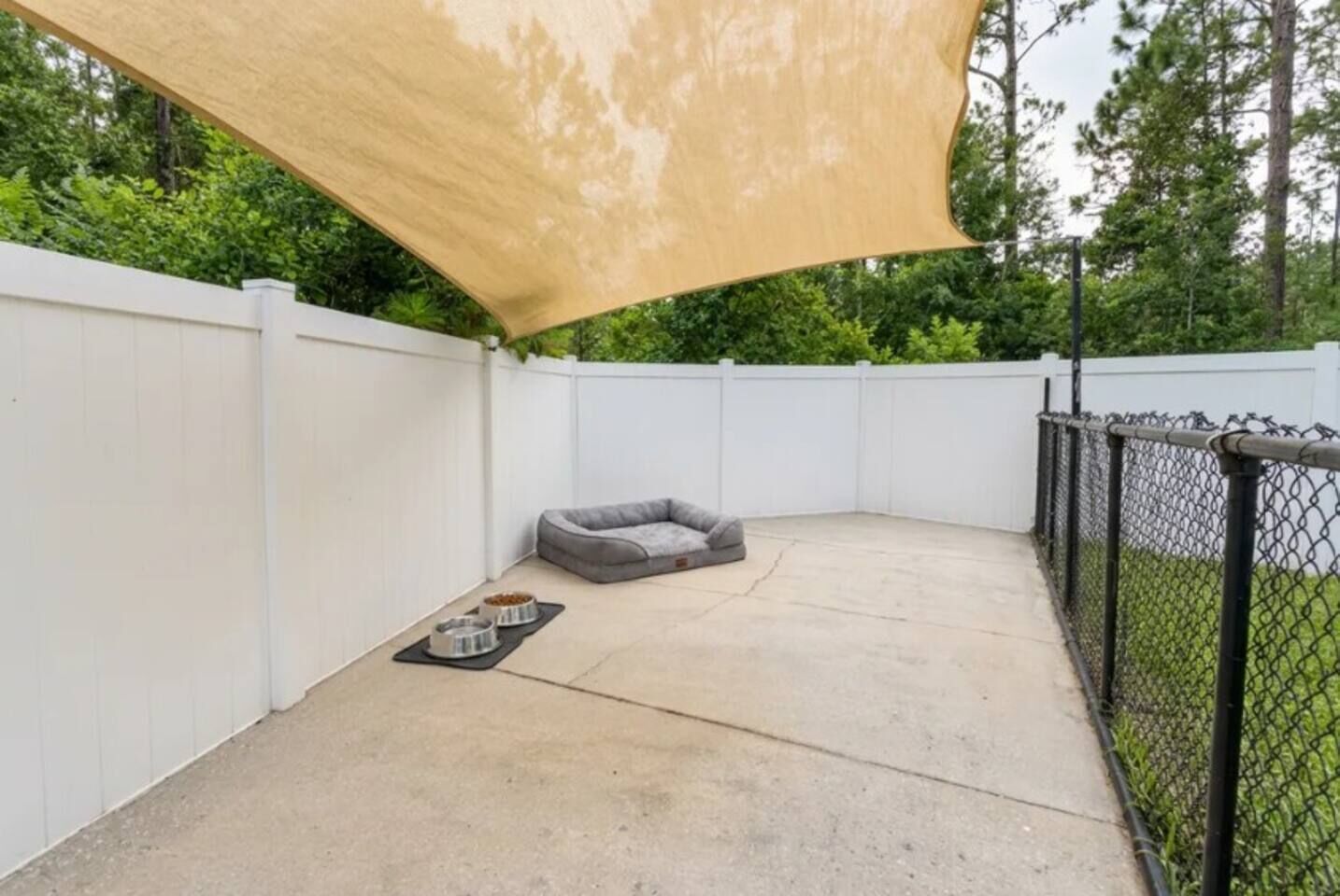 A backyard with a concrete patio, a grey dog bed, two metal bowls, a white fence, and a chain-link fence.