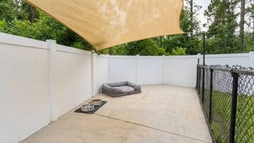 A backyard with a concrete patio, a grey dog bed, two metal bowls, a white fence, and a chain-link fence.