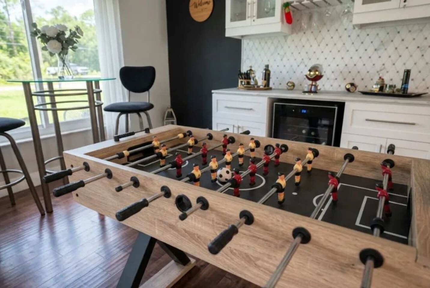 A foosball table in a room with a kitchen in the background, a window with a view of greenery, and a bar area with stools.