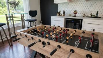 A foosball table in a room with a kitchen in the background, a window with a view of greenery, and a bar area with stools.