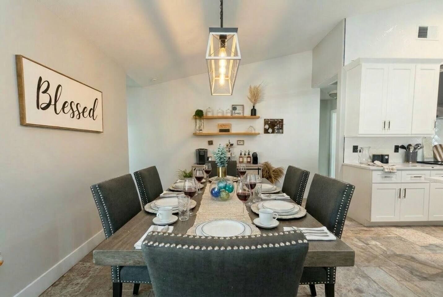 A dining room with a wooden table set for four, surrounded by upholstered chairs. A pendant light hangs above the table, and a 'Blessed' sign is visible on the wall.