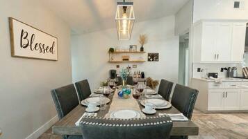 A dining room with a wooden table set for four, surrounded by upholstered chairs. A pendant light hangs above the table, and a 'Blessed' sign is visible on the wall.