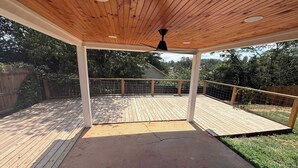 Um deck coberto ao ar livre, com teto de madeira, ventilador de teto preto e guarda-corpo de metal.