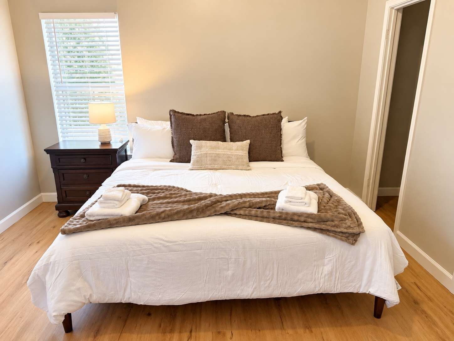 A neatly made bed with white linens and a brown throw blanket, a nightstand with a lamp, and a window with blinds.