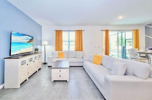 A modern living room with a flat-screen TV, white sofas, a coffee table, and a dining area with a table and chairs. - 8BR Near Disney | Private Pool & Hot Tub (Kissimmee)