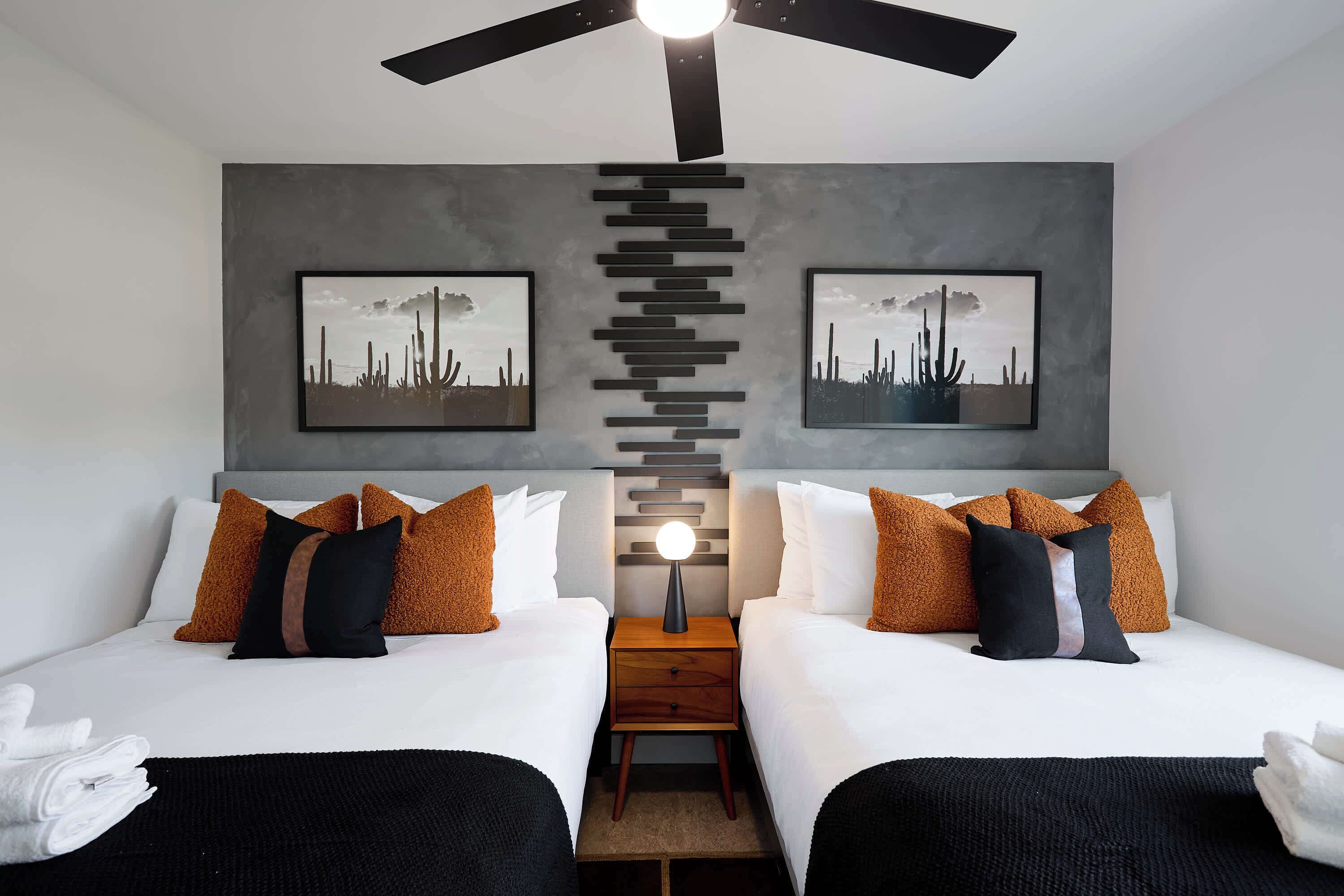 A double room with two beds, a ceiling fan, a nightstand, and artwork on the walls.