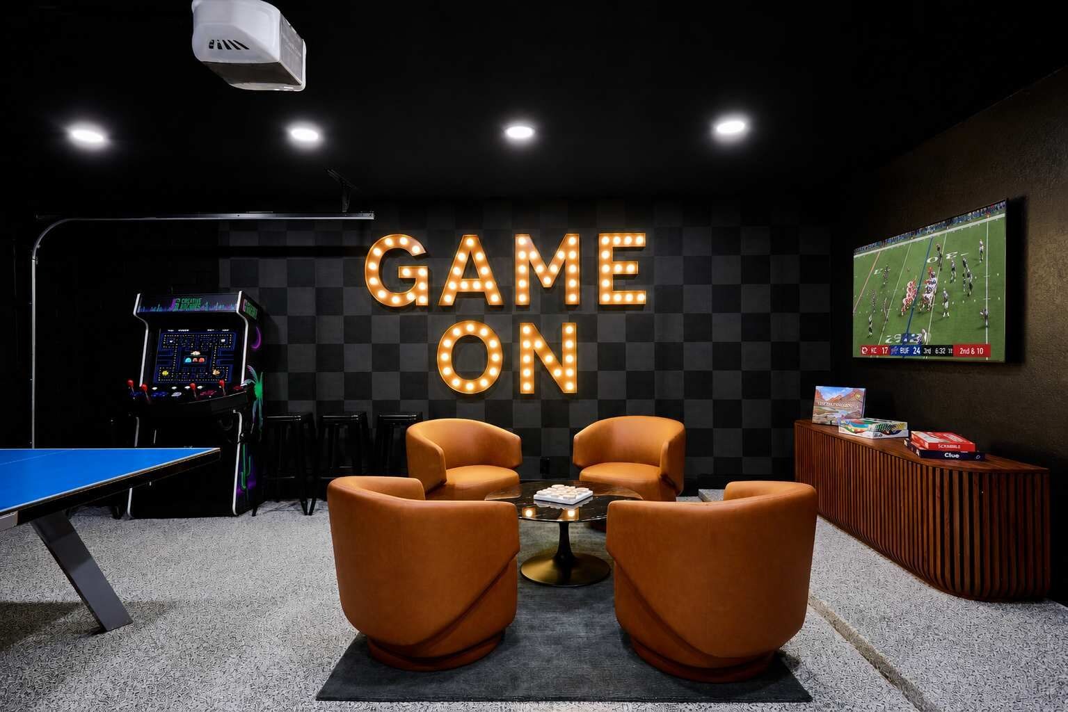 A gaming room with a ping pong table, two armchairs, a coffee table, a gaming console, and a TV displaying a football game.