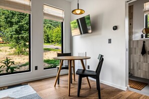 A modern interior with a dining area, a TV mounted on the wall, and large windows offering a view of greenery outside. - Hodo Springs — Birch | A-Frame | Private Sauna, Hot Tub | River Access | Smokies (Sevierville)