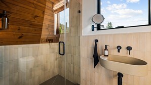 A bathroom with a wooden ceiling, a large window with a view, a round mirror, a sink, and a towel rack. - Hodo Springs — Maple | Luxury A-Frame | Private Sauna, Hot Tub | River | Smokies (Sevierville)