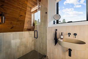 A bathroom with a wooden ceiling, a large window with a view, a round mirror, a sink, and a towel rack. - Hodo Springs — Maple | Luxury A-Frame | Private Sauna, Hot Tub | River | Smokies (Sevierville)