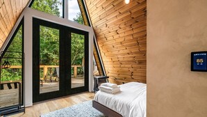 A bedroom with a bed, a digital thermostat, and a view of the outdoors. - Hodo Springs — Maple | Luxury A-Frame | Private Sauna, Hot Tub | River | Smokies (Sevierville)