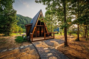 A small, triangular wooden cabin with a sloped roof, a porch, and a gravel pathway leading to it. - Hodo Springs — Pine | A-Frame | Forest Sauna Path + Hot Tub | Smokies (Sevierville)