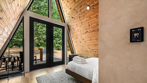 Quarto com teto inclinado de madeira, cama com roupa de cama branca e termostato na parede.