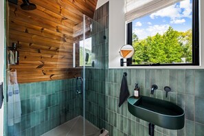 Een moderne badkamer met een houten plafond, een groot raam met uitzicht op bomen, een ronde spiegel en een groene wastafel.