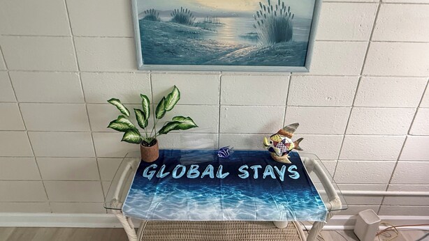 Ein gerahmtes Gemälde einer Küstenlandschaft über einem Tisch mit einer 'GLOBAL STAYS' Unterlage, einem Blumentopf und einer Fischfigur.