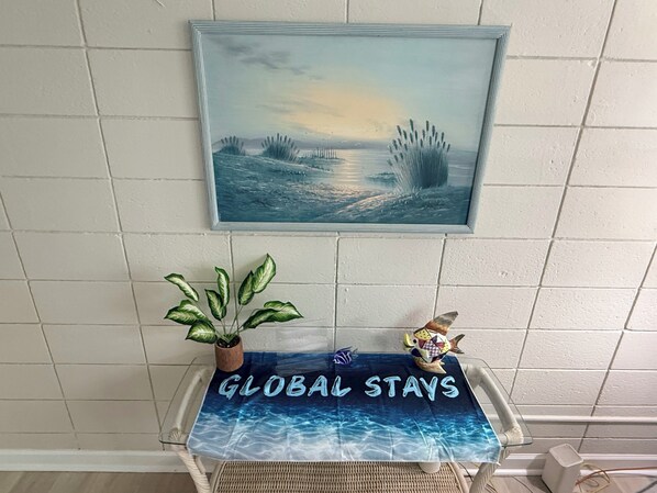 Un tableau encadré représentant un paysage côtier, placé au-dessus d’une table ornée d’un set de table « GLOBAL STAYS », d’une plante en pot et d’une figurine de poisson.