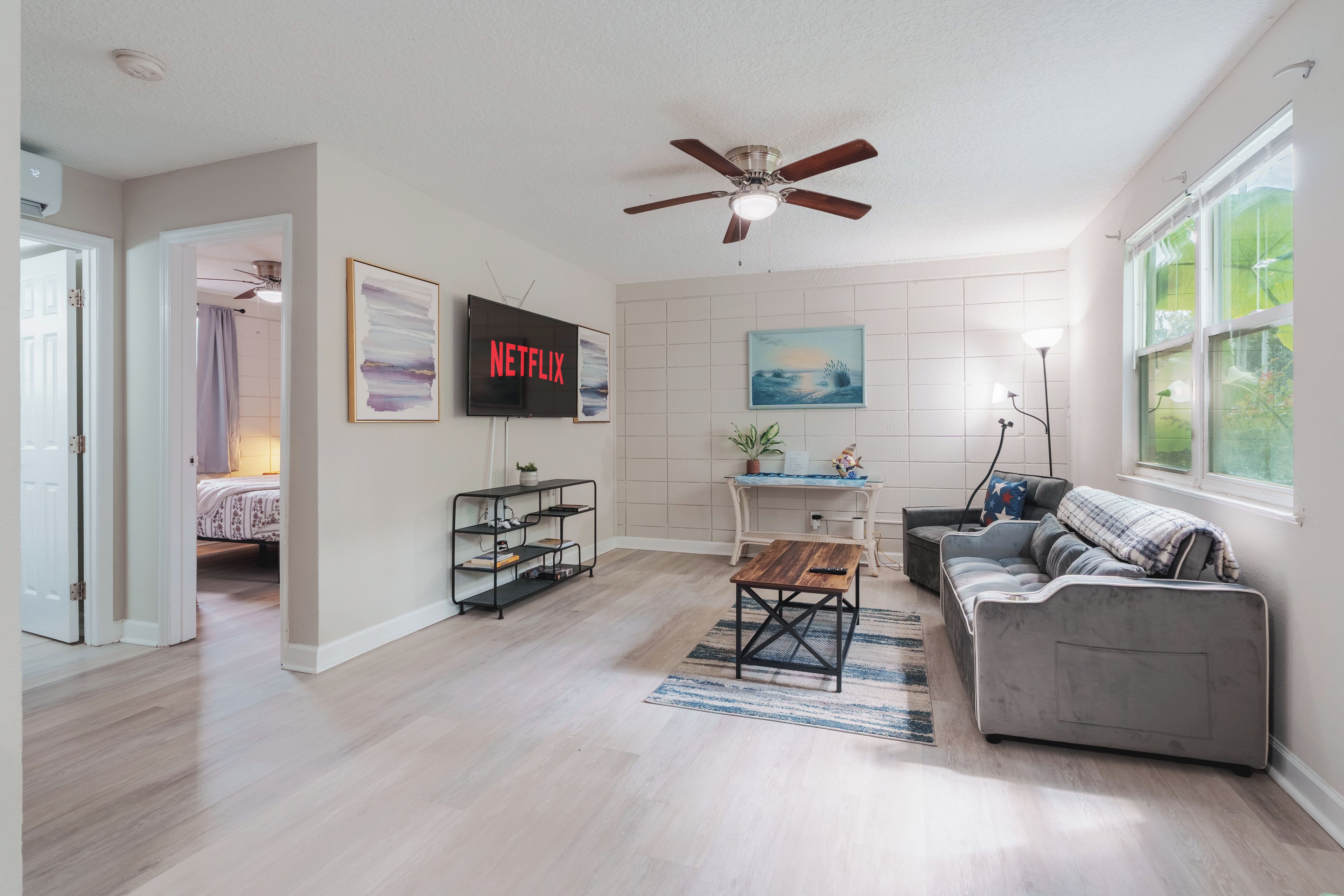A living room with a ceiling fan, a TV mounted on the wall, a bookshelf, a coffee table, a sofa, and a window with a view.