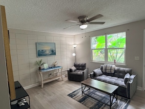 Sala de estar com ventilador de teto, sofá, mesa de centro, mesa lateral com objetos decorativos e janela com vista para área verde.