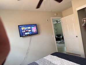 Une chambre à coucher avec un téléviseur fixé au mur qui affiche l’interface d’un service de diffusion en continu, un lit recouvert d’une couvre-lit à motifs et une porte donnant sur une autre pièce.