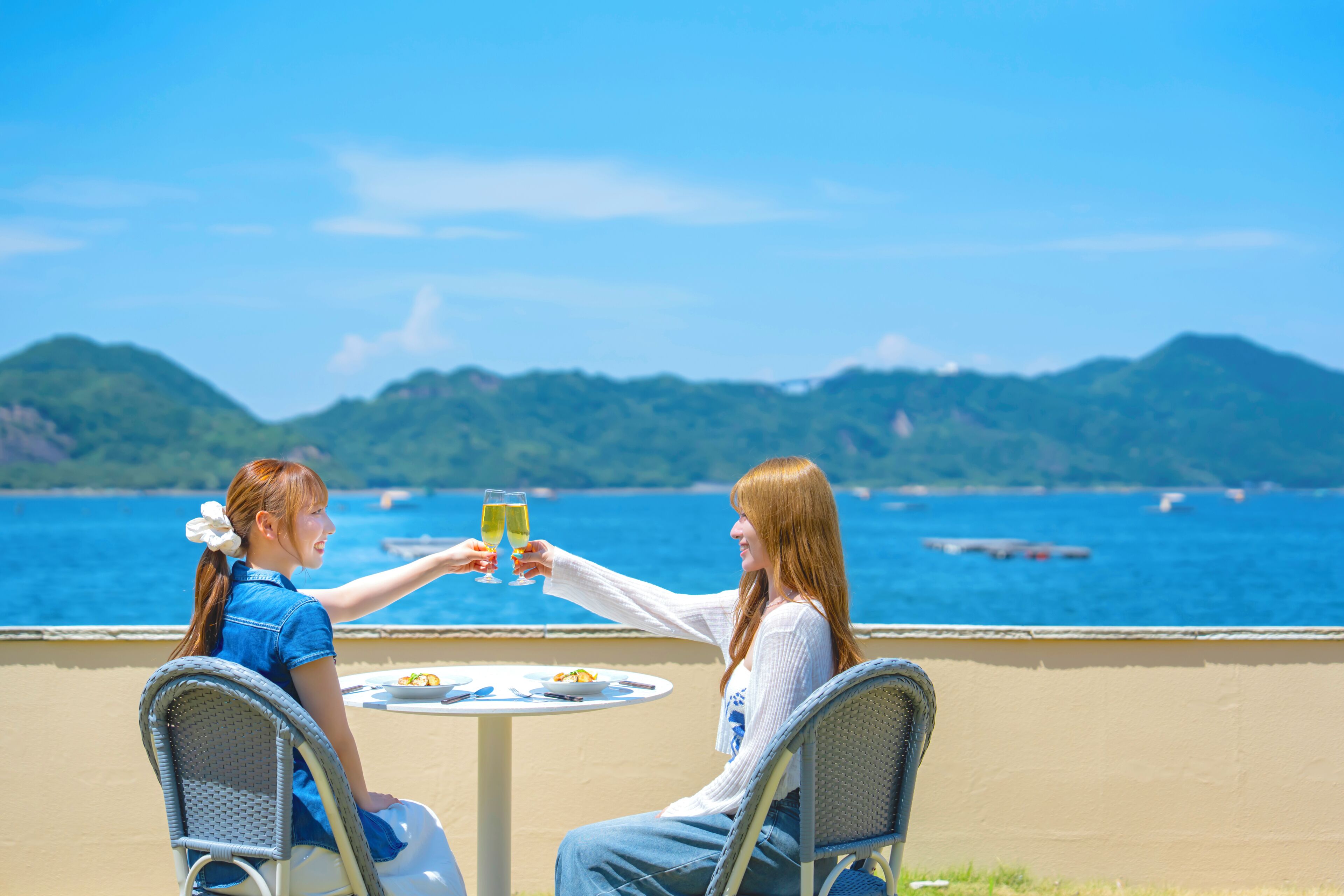 Two people toasting at a table with a view of the sea and mountains in the background.