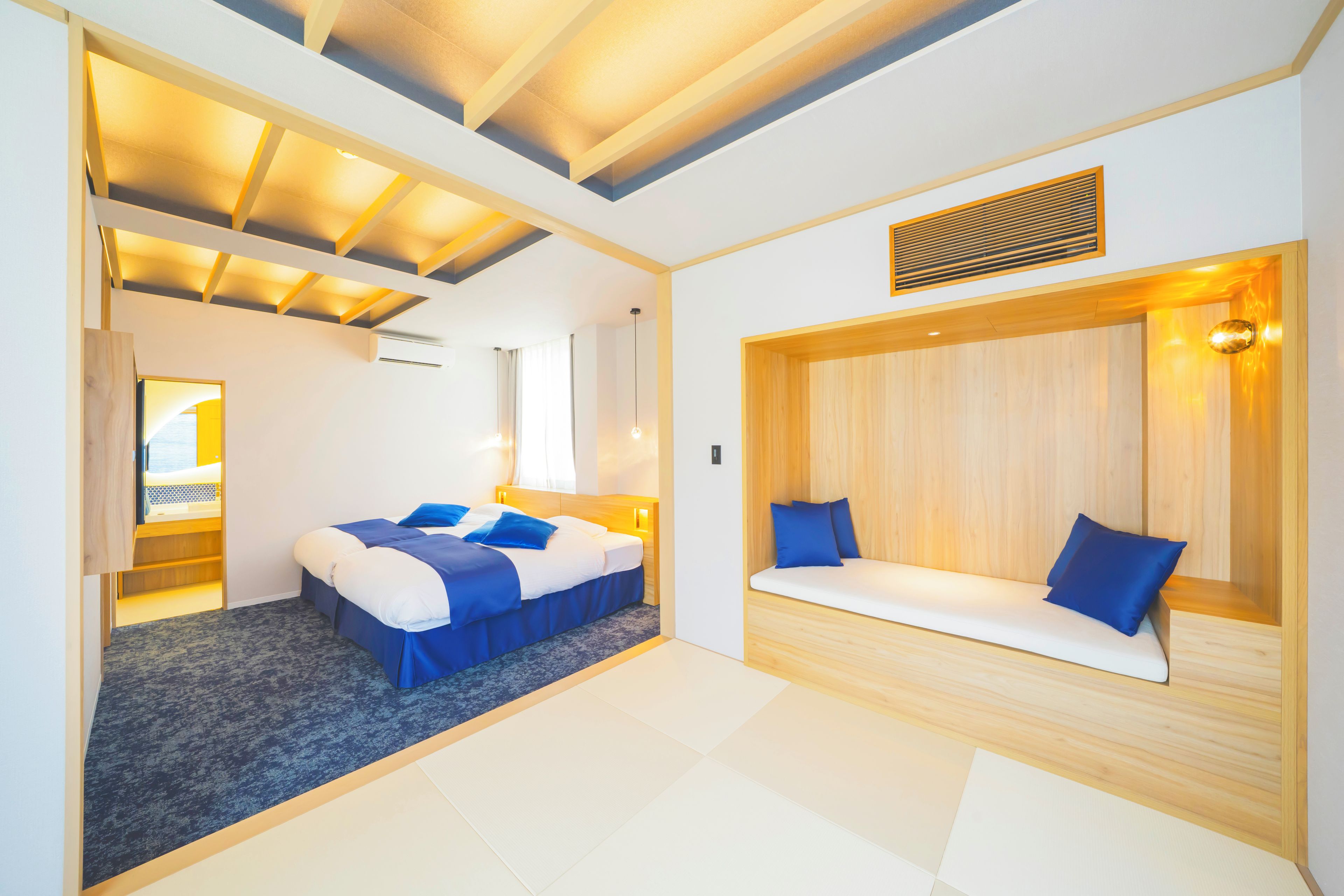 A modern hotel room with a large bed, a built-in bench, and a ceiling with recessed lighting.