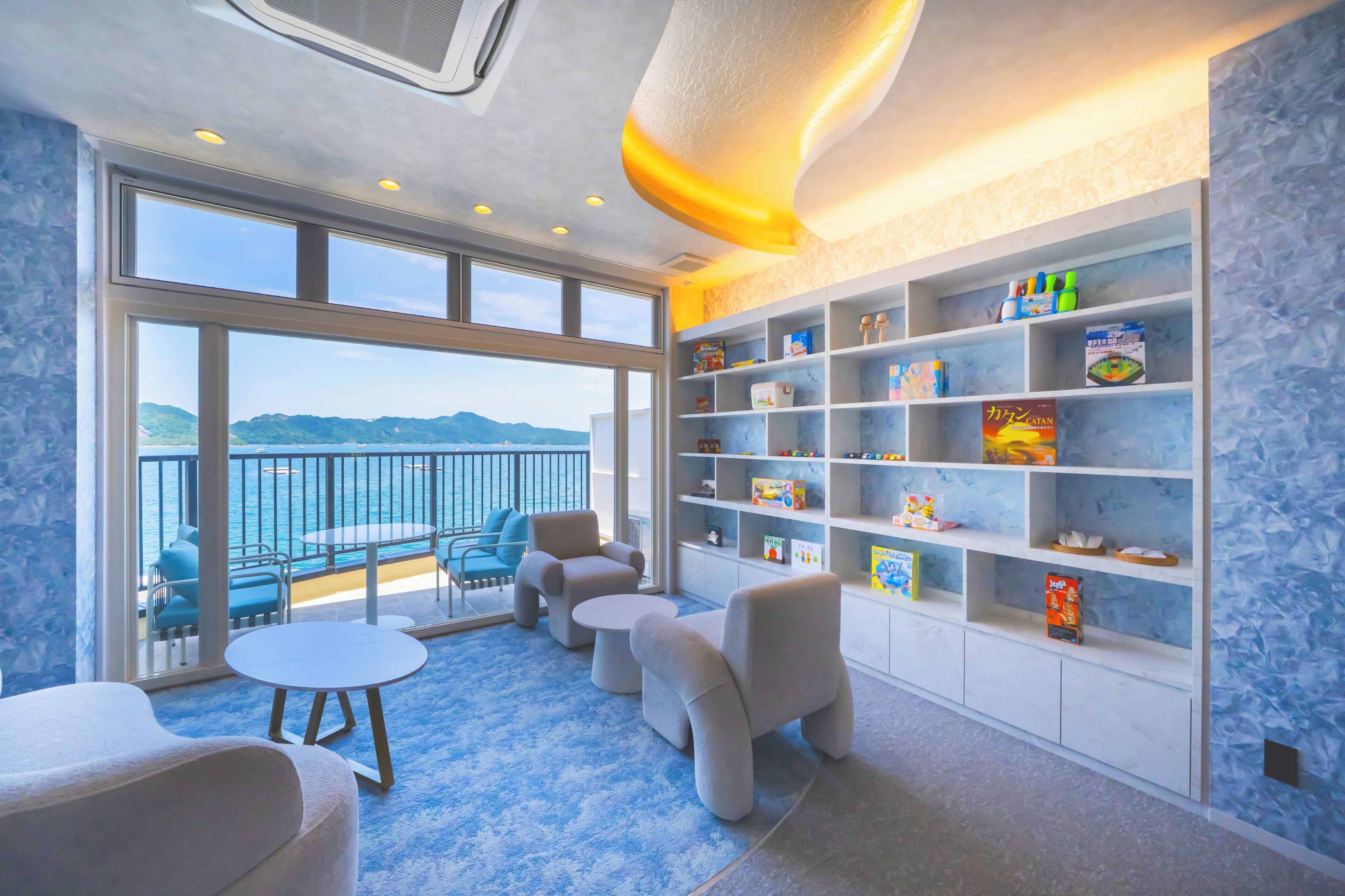A room with a large window overlooking a body of water, a balcony with chairs and a table, and a wall with shelves containing various items.