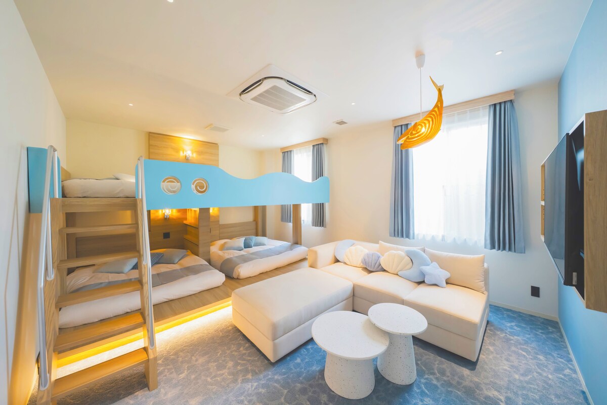 Habitación de hotel moderna con literas, un sofá, dos mesas redondas y un ventanal con cortinas.
