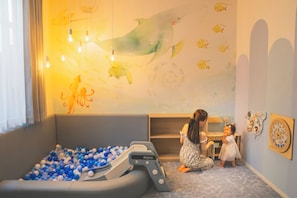 Una sala de juegos con una piscina de pelotas, un niño jugando con una cocina de juguete y una pared decorada con calcomanías de peces y pulpos.