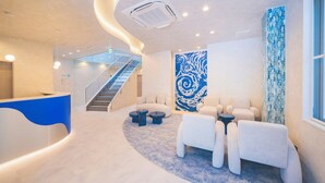 A modern lobby with a curved reception desk, a staircase, a blue abstract wall, and a seating area with white sofas and round tables.