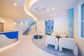 Un vestíbulo moderno con un mostrador de recepción curvo, una escalera, un muro abstracto azul y una zona de estar con sofás blancos y mesas redondas.