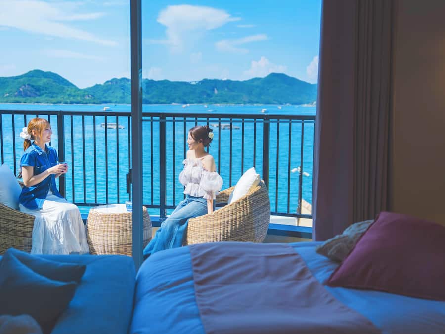 A balcony with wicker furniture, a bed with pillows, and a view of the sea and mountains.