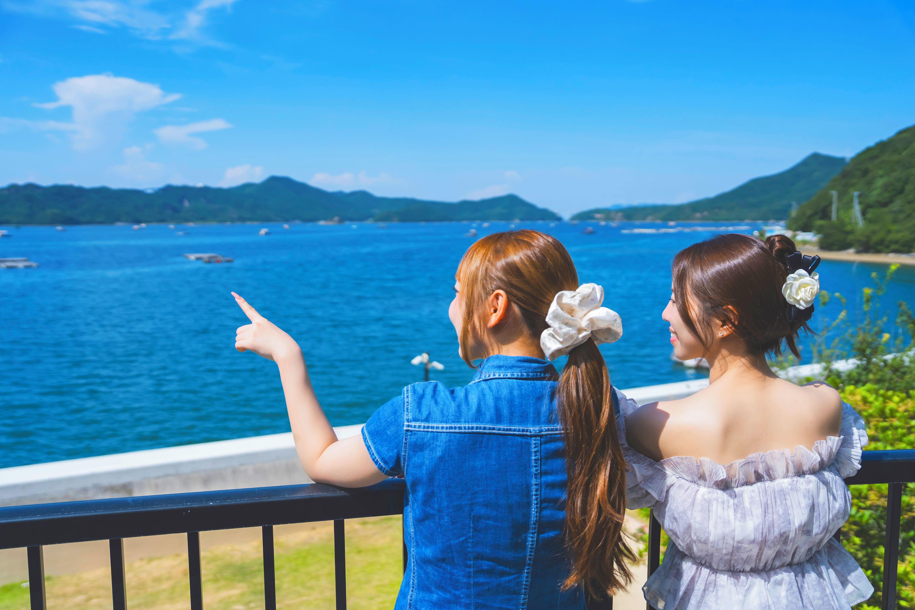 Two individuals overlooking a body of water with boats, a distant shoreline, and hills.