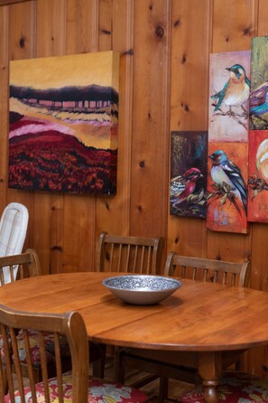 Une table à manger en bois, sur laquelle se trouve un saladier, entourée de chaises, dans une pièce aux murs lambrés et décorée de tableaux.